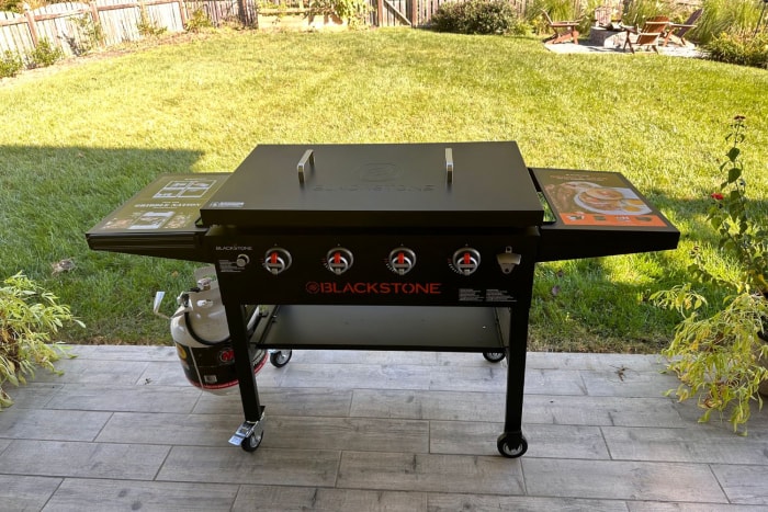 Plaque de cuisson Blackstone de 36 pouces avec couvercle rigide sur patio en pierre dans une cour arrière ensoleillée.