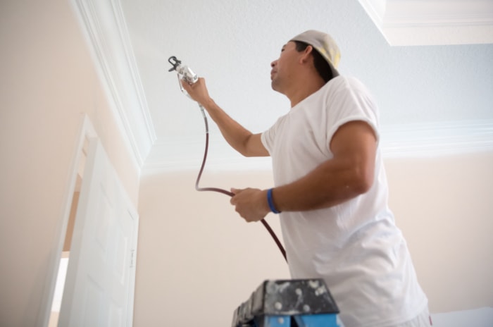 Un peintre en bâtiment utilise un pistolet à peinture pour peindre les moulures du plafond de la maison, debout sur une échelle. Il porte un t-shirt blanc et un pantalon de peintre. Il peint la porte de la pièce.