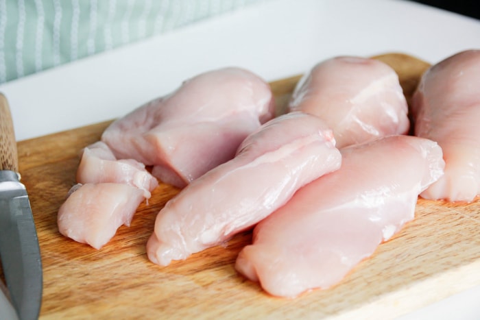 Cinq poitrines de poulet crues sur une planche à découper en bois avec un couteau à proximité.