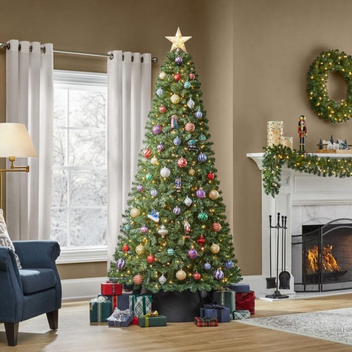 Le sapin festif à LED Home Accents Holiday Prelit est décoré d'ornements et d'une étoile, avec des cadeaux en dessous et une cheminée décorée en arrière-plan.