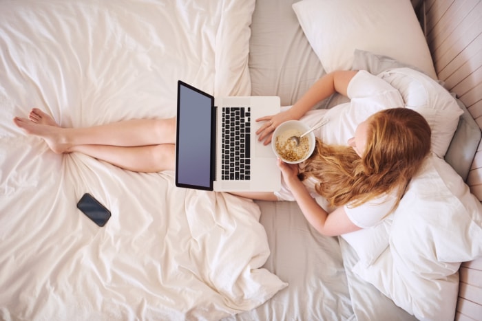 vue aérienne d'une femme au lit avec des draps et une literie blancs, assise avec un ordinateur portable sur les genoux et mangeant un bol de céréales