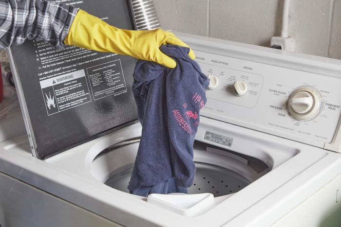 Une femme portant des gants en caoutchouc jaunes place un t-shirt bleu dans une machine à laver à chargement par le haut.