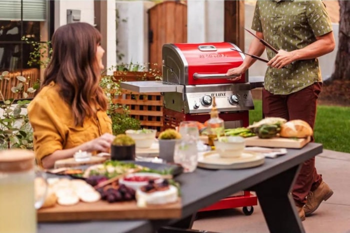 Une femme assise à une table extérieure remplie de nourriture et de boissons tandis qu'un homme se tient debout, tenant des pinces à côté du meilleur barbecue à gaz sous $500