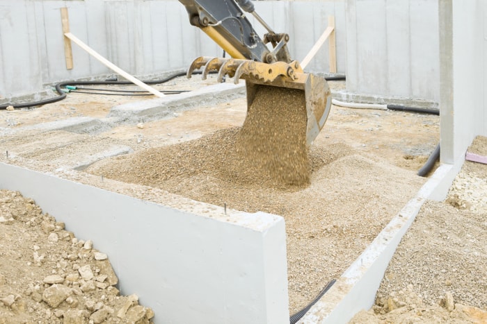 Un godet d'excavatrice déverse des roches à l'intérieur des murs de fondation d'une nouvelle maison. La roche est posée sur la terre et le drain pour un système d'atténuation du radon. Une dalle de béton sera coulée sur la roche pour former le sol.