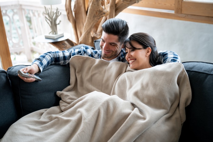 Homme et femme sur un canapé sous une couverture beige, regardant la télévision.