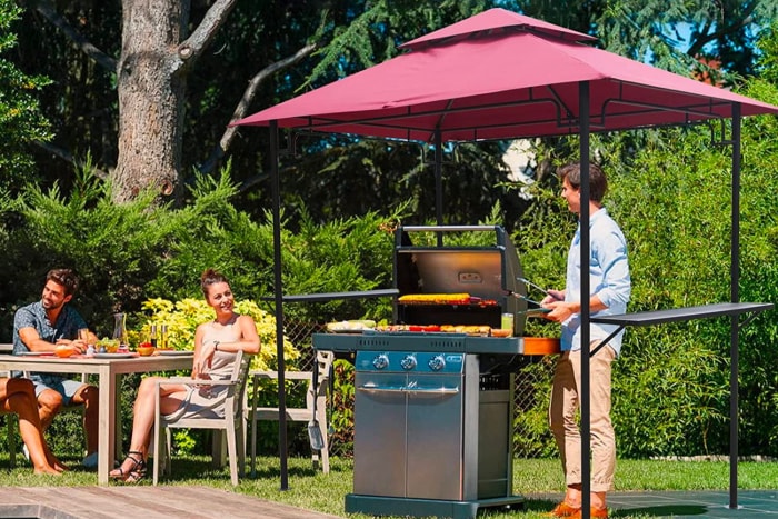 Un homme et une femme à l'extérieur, l'un assis confortablement à une table et l'autre faisant des grillades à l'ombre sous le meilleur gazebo pour grillades.