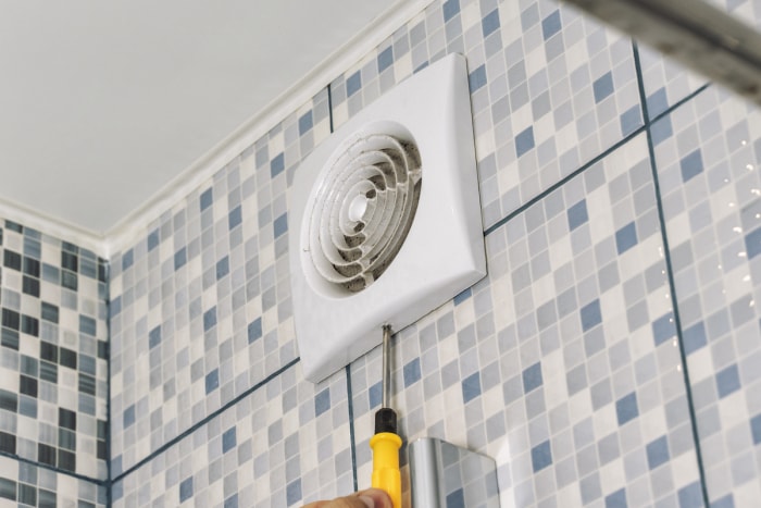 Une personne retire le couvercle du ventilateur d'extraction dans une salle de bain carrelée bleue, grise et blanche.