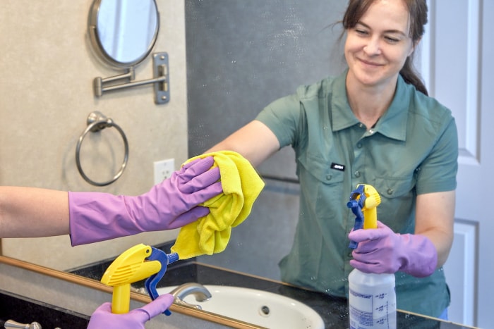 Reflet d'une femme nettoyant un miroir de salle de bain avec un chiffon en microfibre jaune.