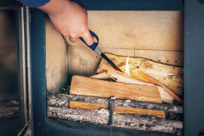 Une main utilise un briquet à long manche pour allumer du bois dans un poêle à bois sur des cendres existantes.