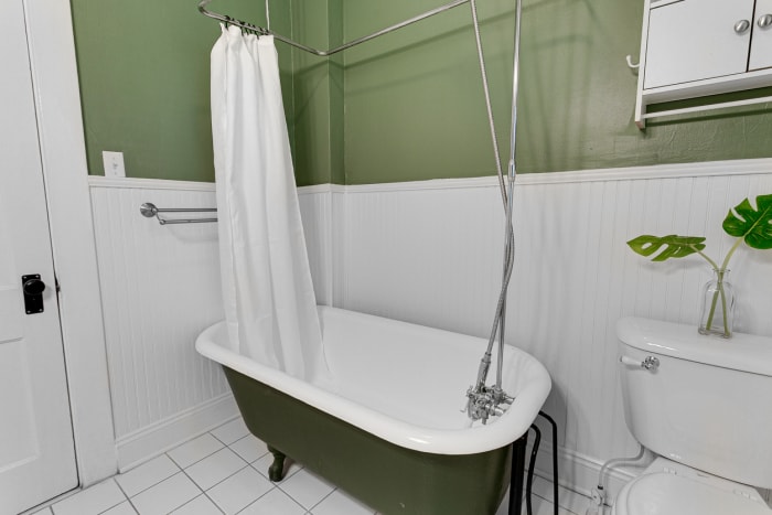 Salle de bains simple et élégante avec murs verts, baignoire blanche, rideau de douche, armoire murale, vase en verre avec des feuilles de monstera, toilettes avec fleur, et design moderne.