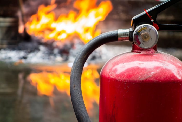 Un extincteur se trouve devant un feu en arrière-plan qui brûle en arrière-plan et se reflète sur le sol.