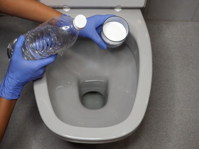 bicarbonate de soude et vinaigre mains gantées tenant les ingrédients au-dessus des toilettes