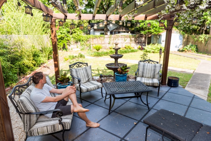 Homme assis sous une pergola dans le jardin avec des meubles de patio foncés et une fontaine.