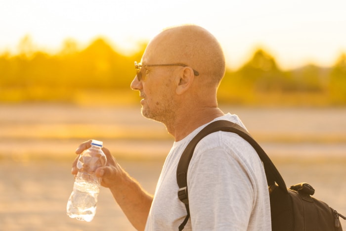 Un homme chauve portant des lunettes de soleil boit dans une bouteille d'eau en plastique tandis que le soleil se couche en arrière-plan.