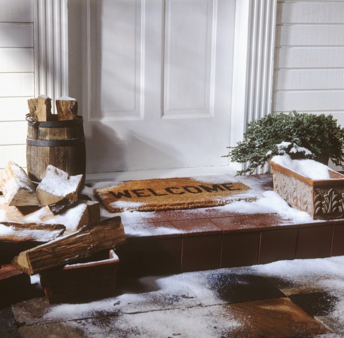 Porche d'entrée en hiver, avec un paillasson, du bois de chauffage dans des tonneaux et d'autres décorations.