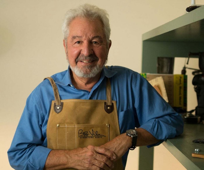 Bob Vila, cheveux et barbe grisonnants, sourit en portant une salopette dans un atelier.