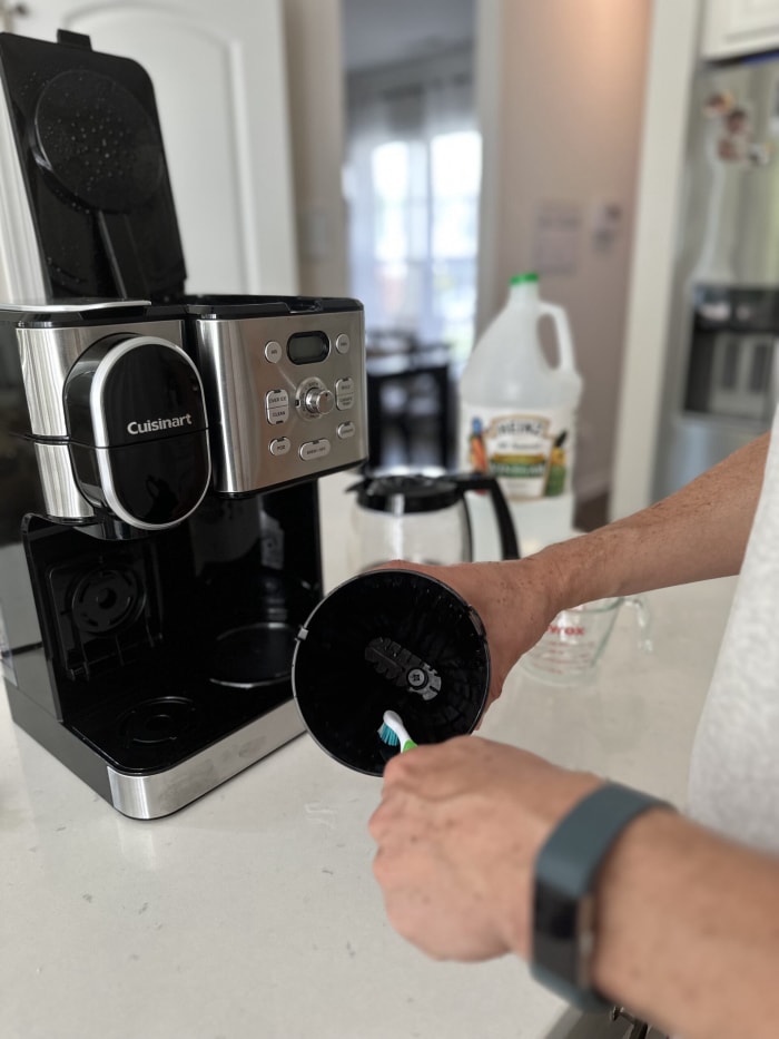 utiliser une brosse à dents pour nettoyer le panier de la cafetière avec la cafetière posée sur le comptoir de la cuisine