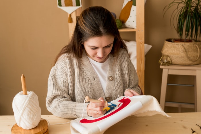 iStock-1395018063 decorate with crafts Femme brodant à la main image arc-en-ciel sur toile. Jeune femme enfonçant l'aiguille à poinçonner dans le tissu de base dans un atelier.