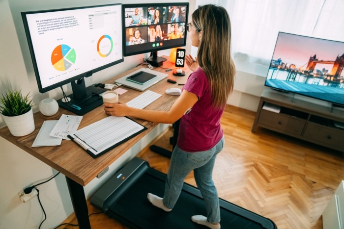 Femme dans un bureau à domicile avec deux écrans, un bureau avec tapis de marche, une télévision et un téléphone.