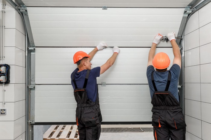 Deux ouvriers portant des casques de sécurité orange installent une porte de garage.