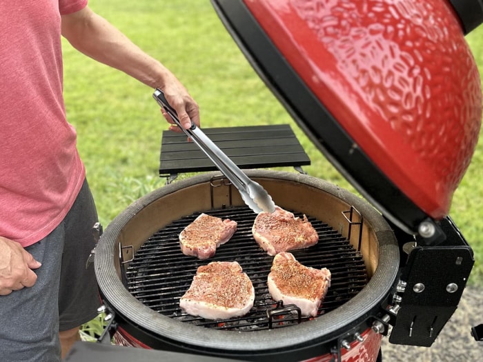 Le gril Kamado Joe Classic Joe ouvert pendant qu'une personne utilise des pinces pour retourner la viande en train de cuire