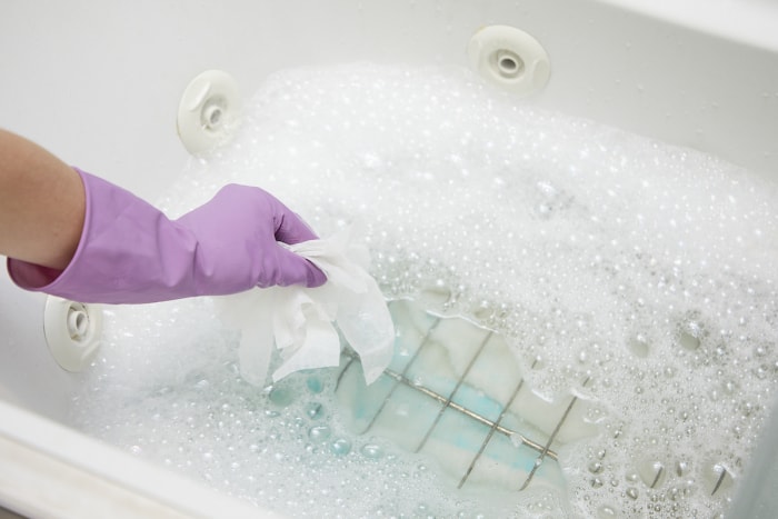 Une femme portant des gants en caoutchouc utilise des feuilles assouplissantes et du savon à vaisselle pour nettoyer les grilles du four dans une baignoire.