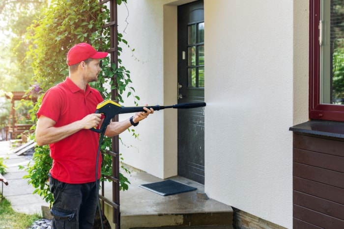 Un homme en chemise rouge et chapeau nettoie une maison à haute pression.