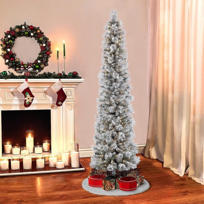 L'arbre à crayons floqué et pré-éclairé de Puleo International avec des cadeaux en dessous est installé à côté d'une cheminée remplie de bougies allumées et décorée d'une couronne et de bas.