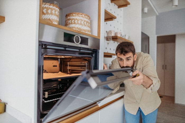 un homme barbu en chemise tannée tient un tournevis pendant qu'il regarde à l'intérieur d'une cuisinière murale dans une cuisine blanche