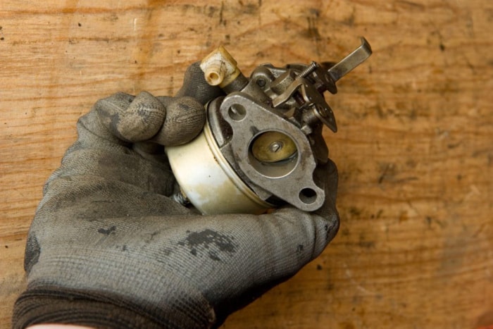 Une main gantée tient un carburateur de petit moteur sale sur un fond en bois.