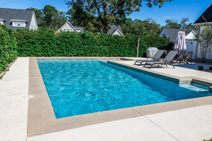 Une nouvelle piscine rectangulaire avec des bords en béton beige dans l'arrière-cour clôturée d'une maison de nouvelle construction avec des haies d'intimité.