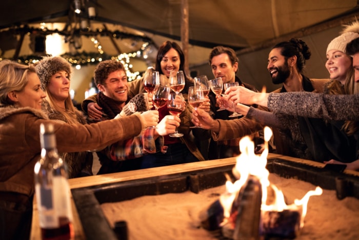 Un groupe de personnes tiennent des verres à vin autour d'un foyer sous un grand auvent.