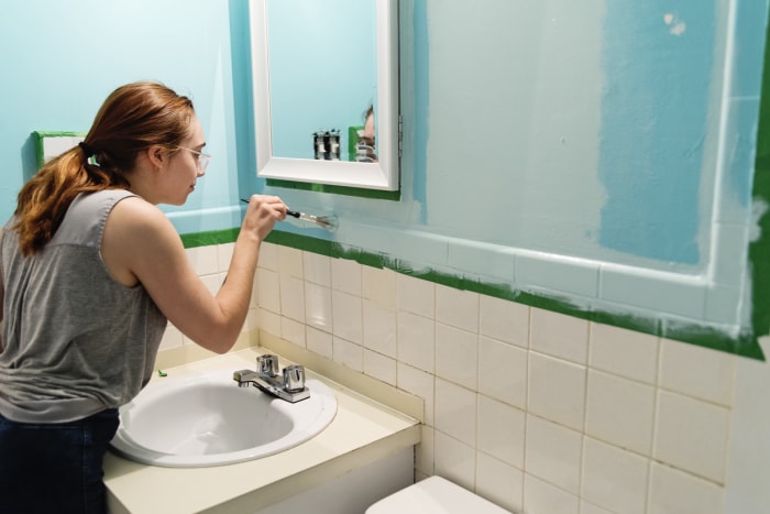 Une jeune femme peint les murs de la salle de bain en bleu vif avec un robinet autour du miroir et du comptoir de la vanité.