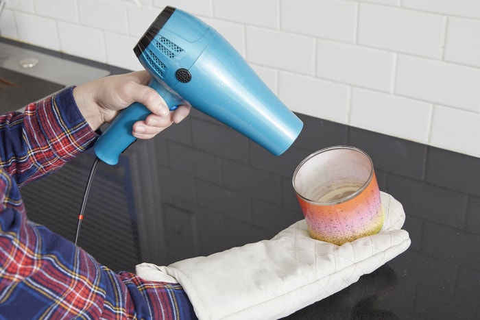 Femme portant un gant de cuisine, utilisant un sèche-cheveux pour faire fondre de la cire de bougie.