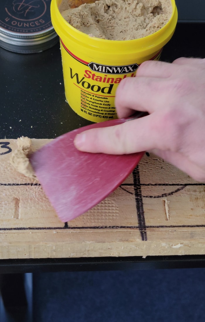 Une personne utilisant un couteau à mastic pour gratter l’excédent de remplissage pour bois teintable Minwax d’une planche de bois récemment remplie.