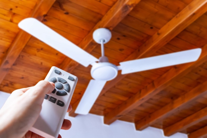 Gros plan d'une main utilisant une télécommande pour faire fonctionner un ventilateur de plafond monté dans une maison sur un plafond en bois.