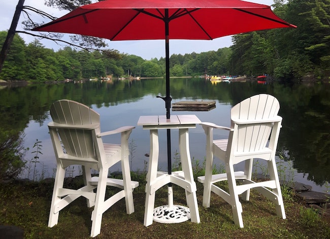 Deux grandes chaises Adirondack blanches avec une petite table et un parasol rouge devant le lac