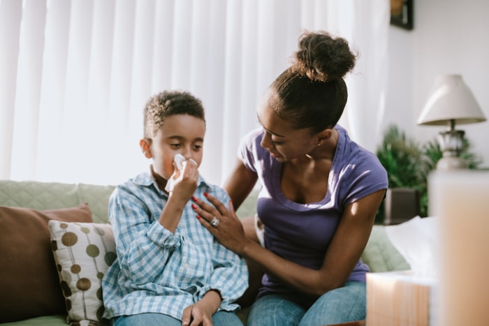 une jeune mère réconforte son fils qui a un mouchoir sur le nez pendant qu'ils sont assis sur un canapé beige avec une lampe en arrière-plan