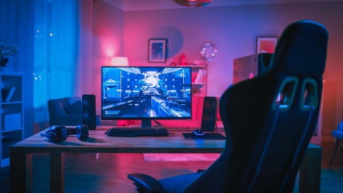 Puissant ordinateur personnel pour gamer avec jeu de tir à la première personne à l'écran. Le moniteur est posé sur la table à la maison. Une pièce confortable au design moderne est éclairée par une lumière néon rose.