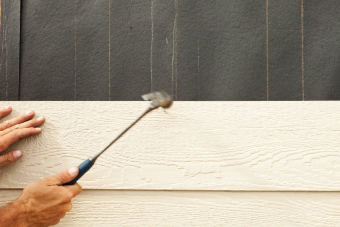 Un ouvrier du bâtiment utilise un marteau pour installer un revêtement sur un mur extérieur d'une maison recouvert de papier goudronné. On observe un flou de mouvement dans le marteau et la tête du clou est visible à côté de la tête du marteau.