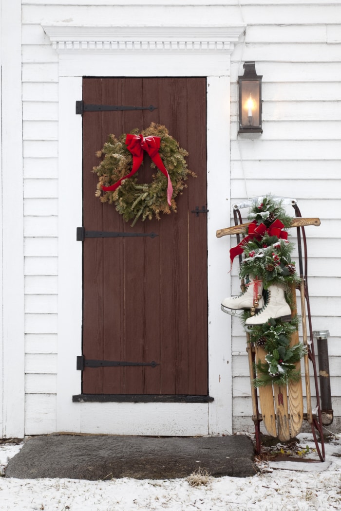 Traîneau et patins antiques appuyés contre le revêtement blanc de la maison, avec une couronne de Noël sur la porte d'entrée marron adjacente.