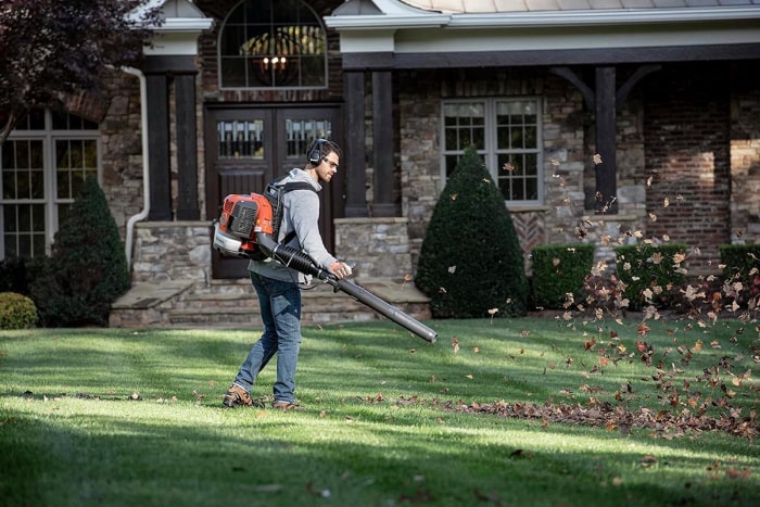 Une personne utilisant un souffleur à dos Husqvarna pour déplacer les feuilles dans une cour avec une grande maison en arrière-plan.