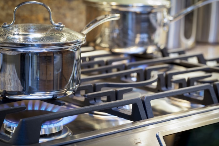 gros plan d'une casserole en acier inoxydable avec poignée et couvercle en verre sur une cuisinière avec du gaz allumé et une autre casserole floue en arrière-plan