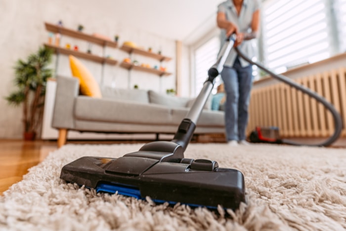 Une femme en jeans utilise un aspirateur pour nettoyer un tapis beige.