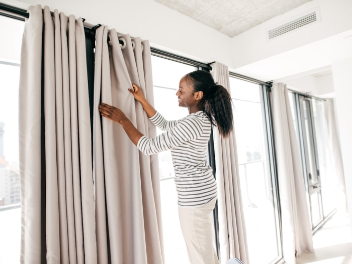 Femme sur un escabeau accrochant des rideaux beiges sur un mur de fenêtres.