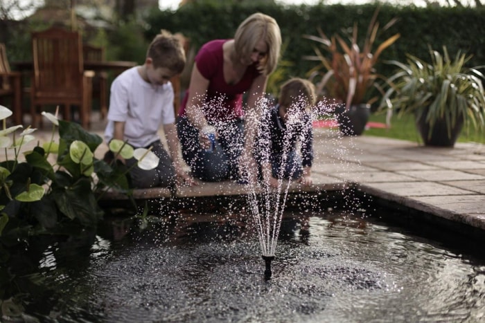 Une femme et deux enfants agenouillés à côté d'un étang avec une fontaine pulvérisant de l'eau