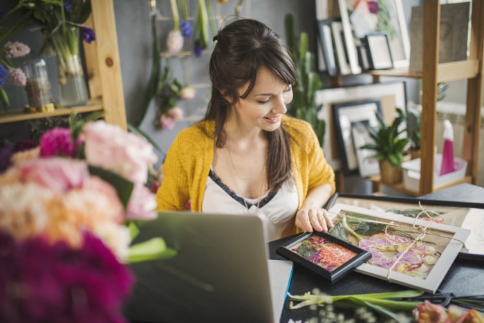 A-young-woman-looks-over-her-framed-floral-photos-surrounded-by-plants-and-shelves.