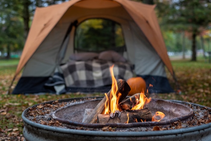 Un foyer brûle devant une tente de camping avec des oreillers et des couvertures à l'intérieur.