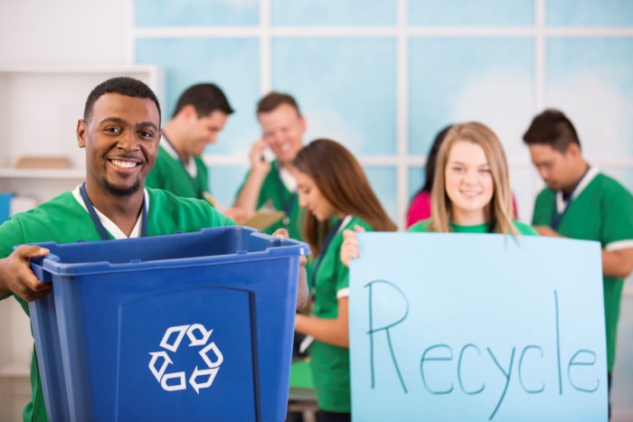 Des bénévoles tiennent un bac de recyclage et une pancarte.