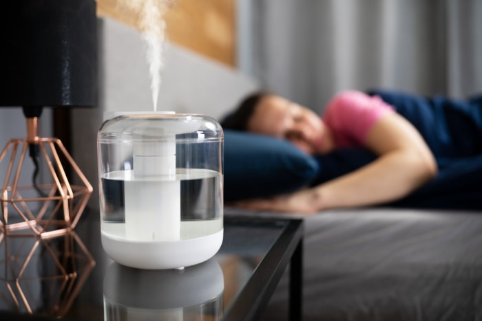 Un humidificateur d'air portable est posé sur une table de chevet à côté d'une lampe noire, tandis qu'une femme en chemise rose dort à l'arrière-plan.